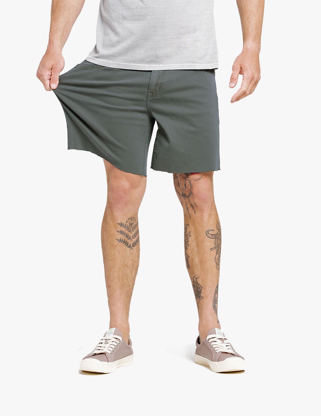 Man wearing gray stretch shorts and beige sneakers, pulling the shorts to show stretchiness, with visible leg tattoos.