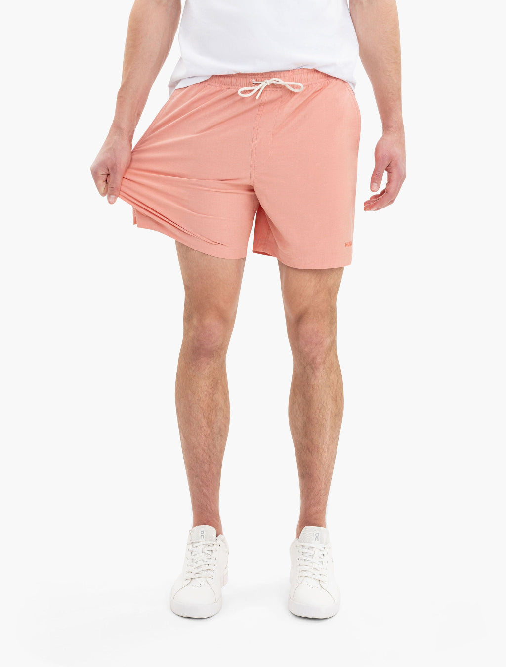 Man wearing light pink shorts stretching the fabric to show its elasticity, paired with white sneakers and a white shirt cropped out of frame.