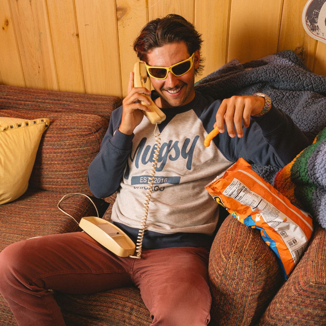 Man on couch wearing yellow sunglasses and 'Mugsy EST 2016' sweatshirt, holding a landline handset and a cheese puff from an open snack bag.