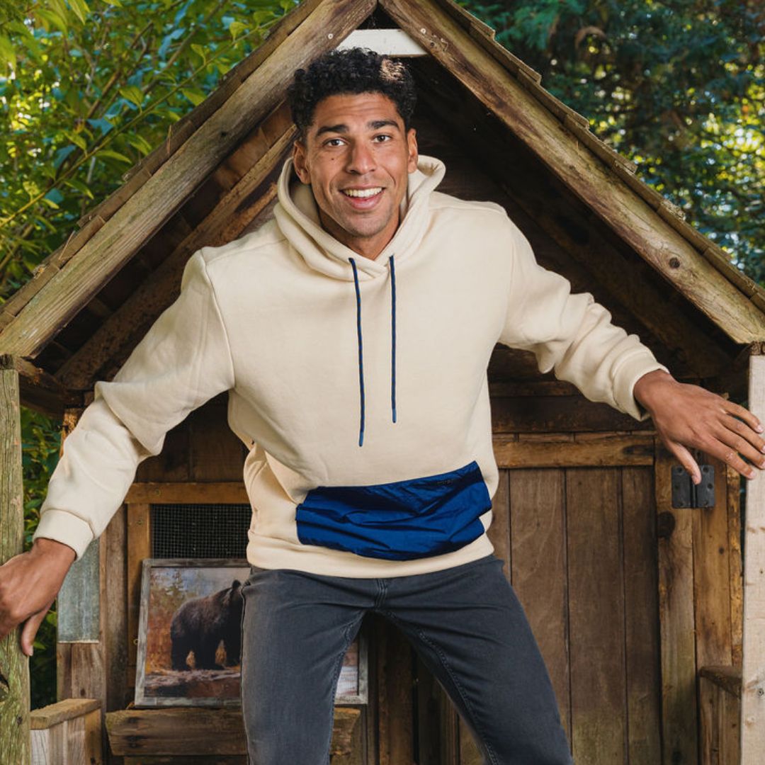 Smiling man in cream hoodie with navy pouch leaning from a small wooden playhouse; framed bear photo visible inside.