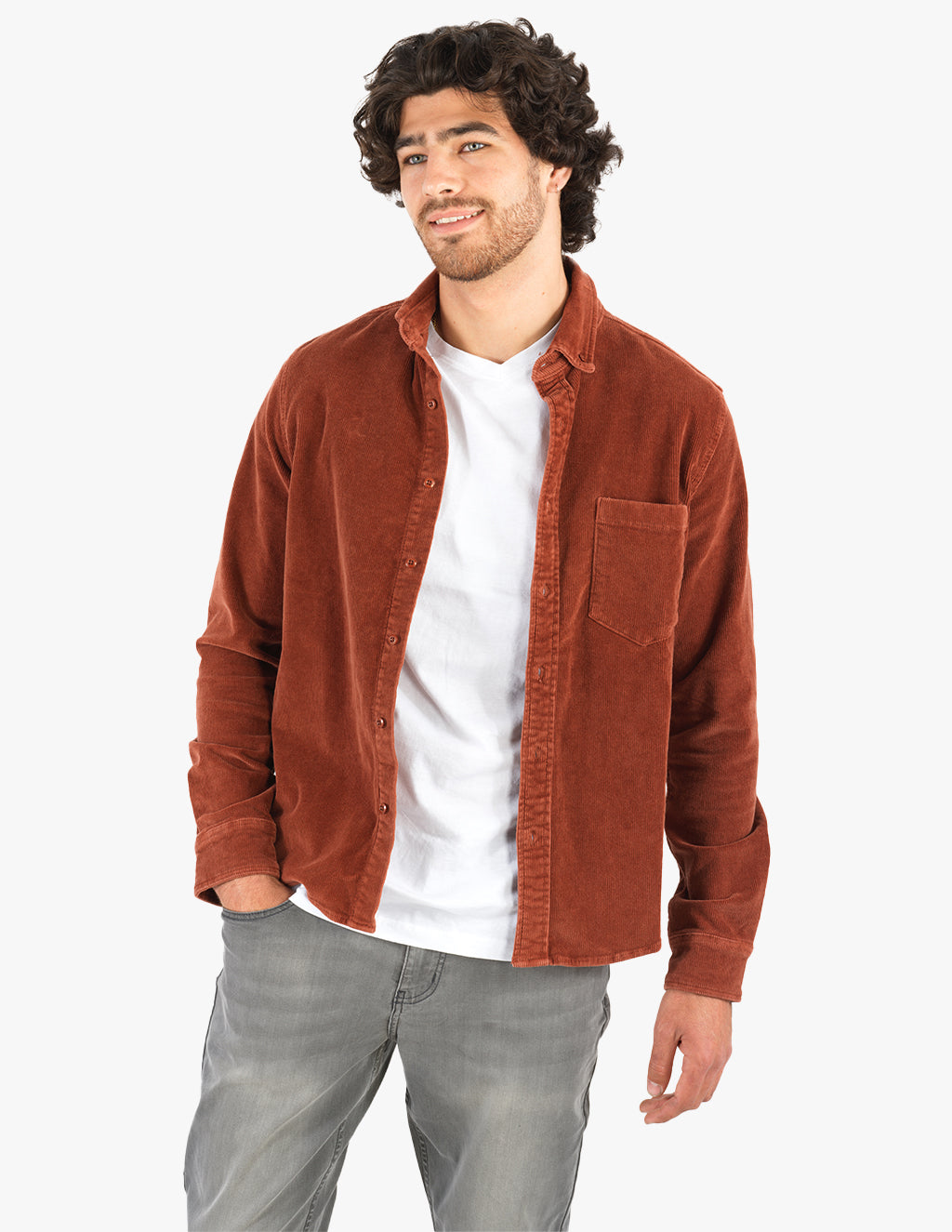 Young man standing with hands in pockets wearing rust corduroy button-up shirt over white T-shirt and gray jeans, smiling