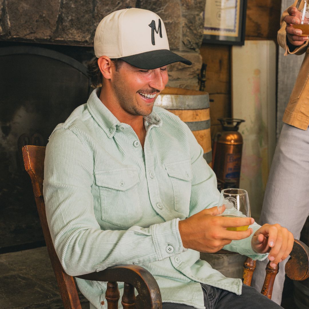 Smiling man in pale green corduroy shirt and baseball cap with 'M', sitting and holding a glass of beer