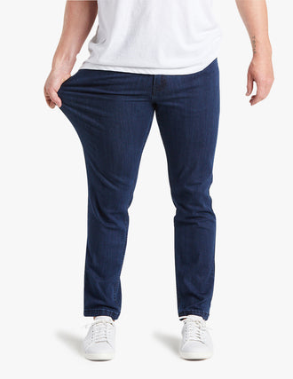 Waist-down view of dark blue slim stretch jeans; left hand stretching fabric; white sneakers; forearm tattoo 'TROIS'.