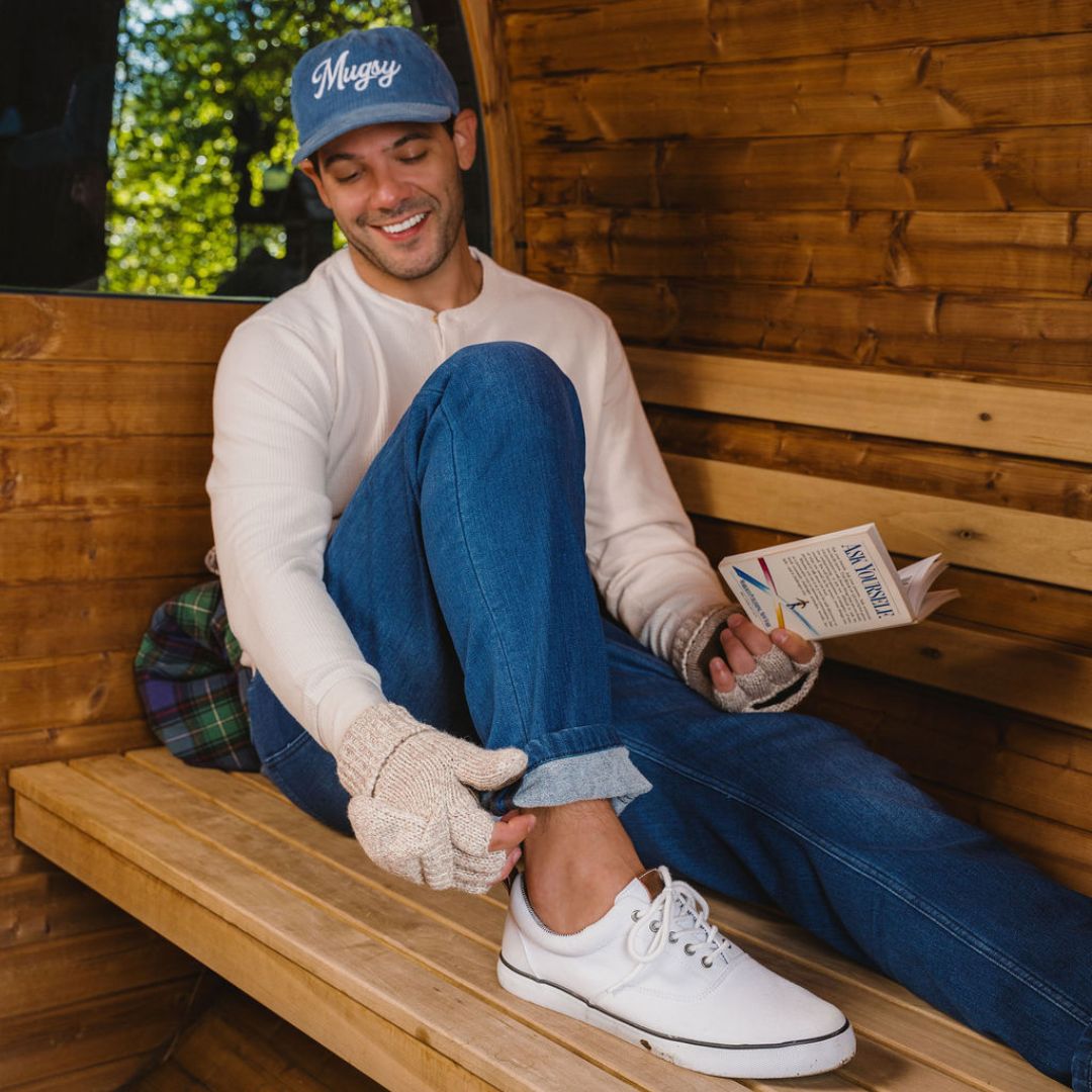 Man on wooden bench reading a book titled 'ASK YOURSELF', wearing a 'Mugsy' cap, fingerless knit gloves, and white sneakers.