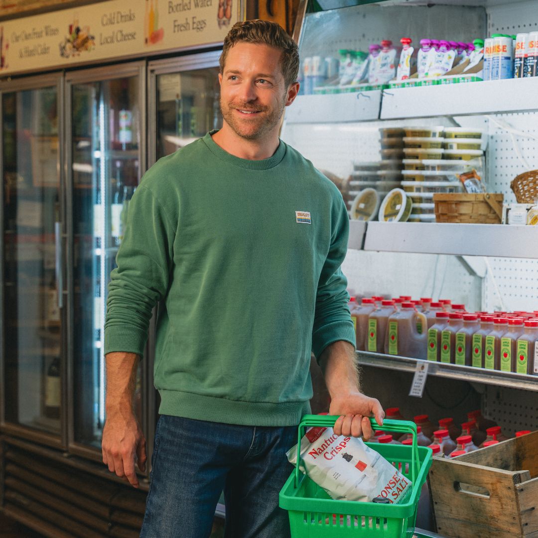 Man in green sweatshirt holding a green shopping basket with a bag labeled 'Nantucket Crisps' and 'ROSEMARY SEA SALT' in a grocery deli aisle.