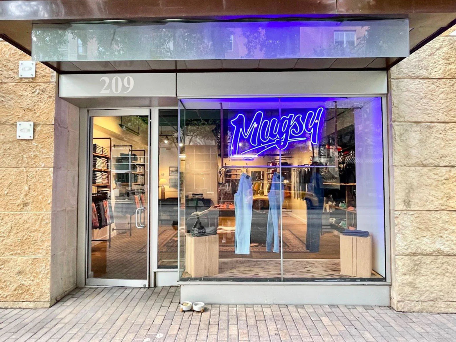 Mugsy storefront at 209 W 2nd St, Austin, TX, featuring a neon sign and clothing display.
