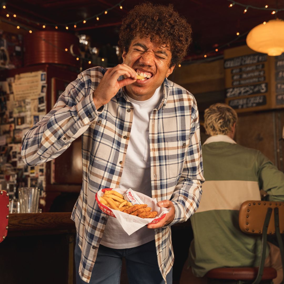 Young man in plaid shirt biting a french fry, holding a basket of fries and chicken strips at a diner counter.