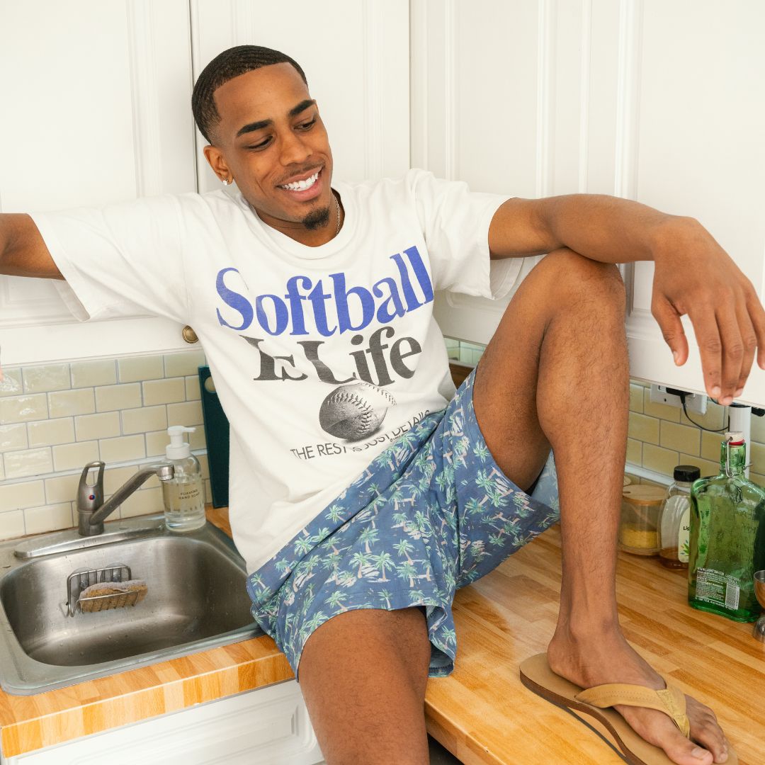 Smiling young man on a kitchen counter wearing a white t-shirt reading: Softball Is Life THE REST IS JUST DETAILS.