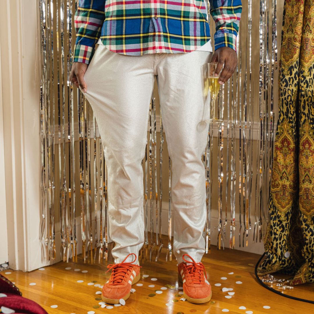 Waist-down view: metallic silver pants and plaid shirt, holding champagne flute; orange sneakers and confetti on floor.