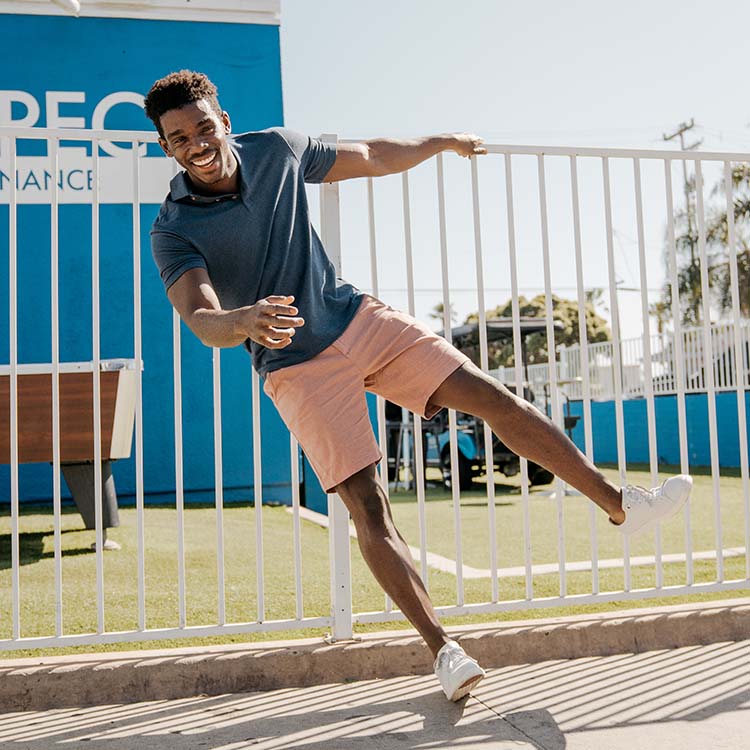 A man wearing a blue polo shirt and pink shorts, smiling and leaning against a fence.