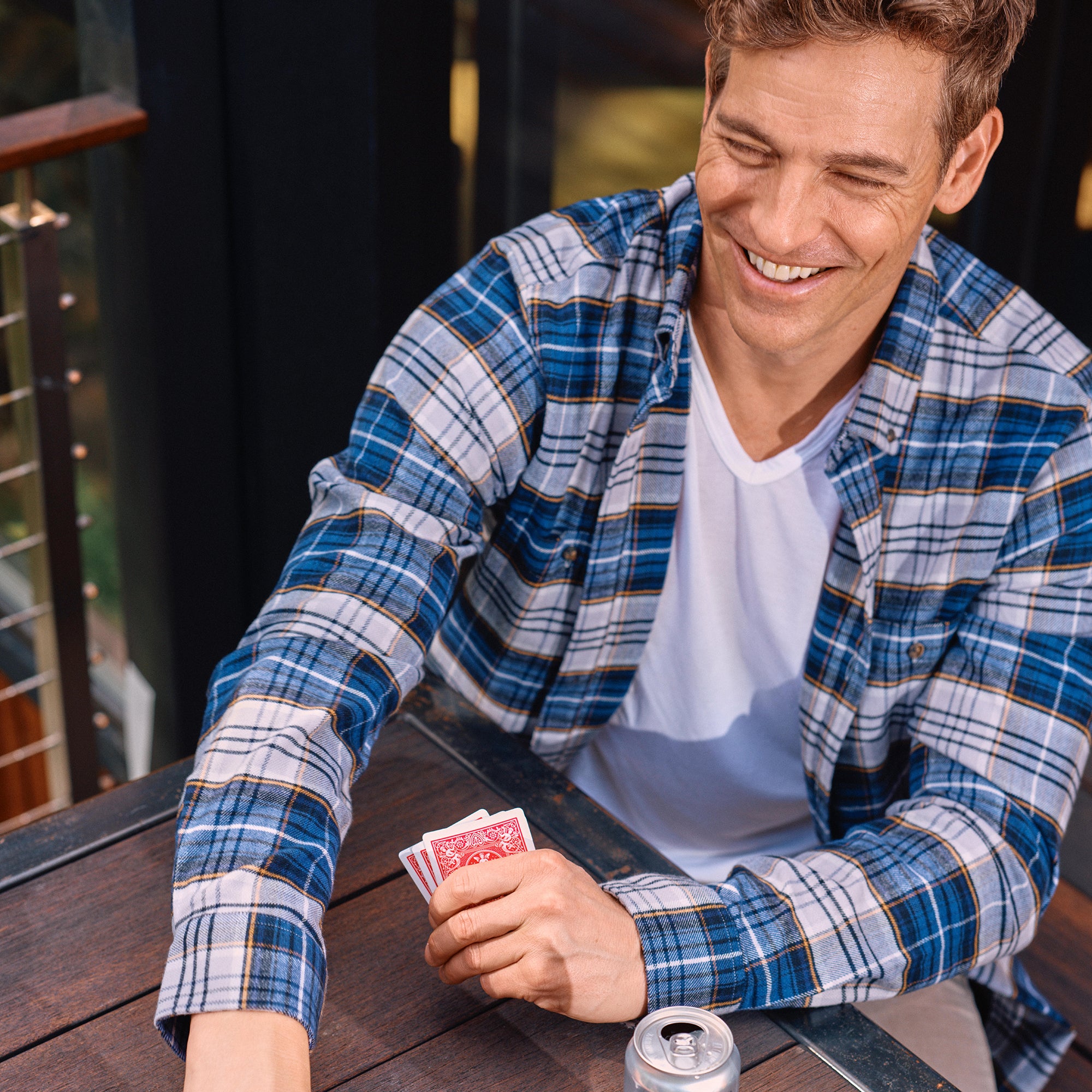 Man in a plaid shirt holding playing cards at a table with a drink.