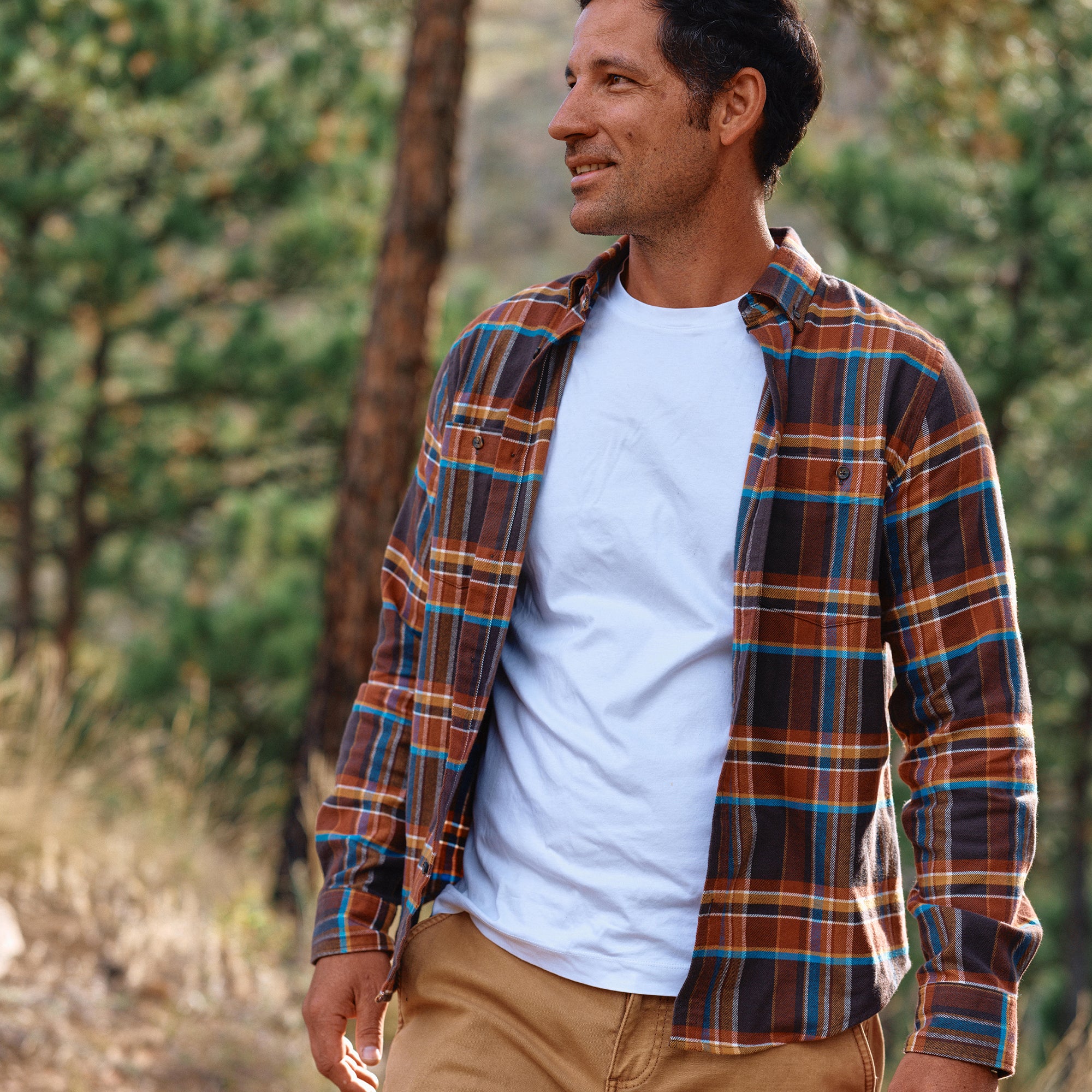 Man wearing a plaid shirt over a white t-shirt in a forest setting.