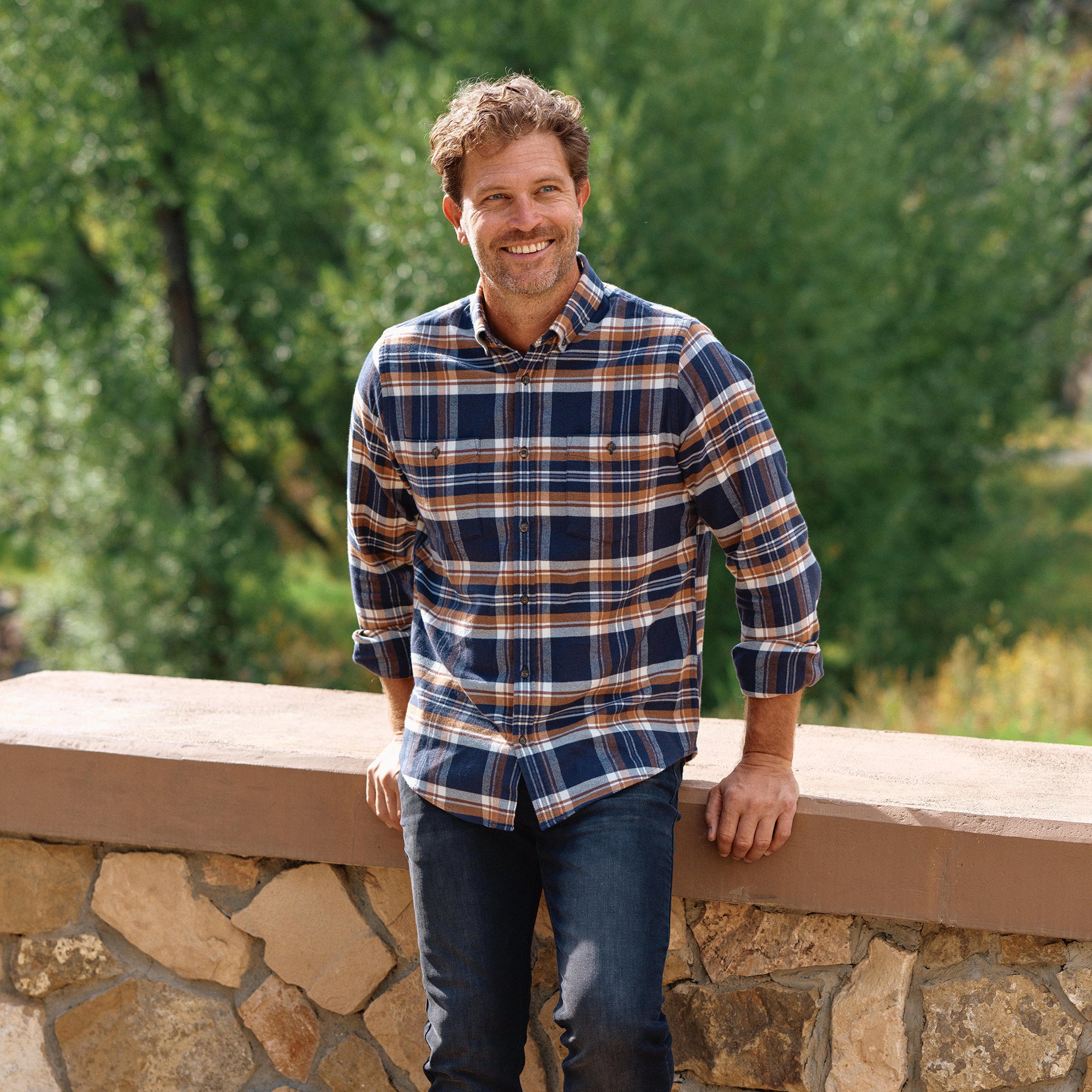 Man wearing a plaid shirt standing outdoors by a stone wall.