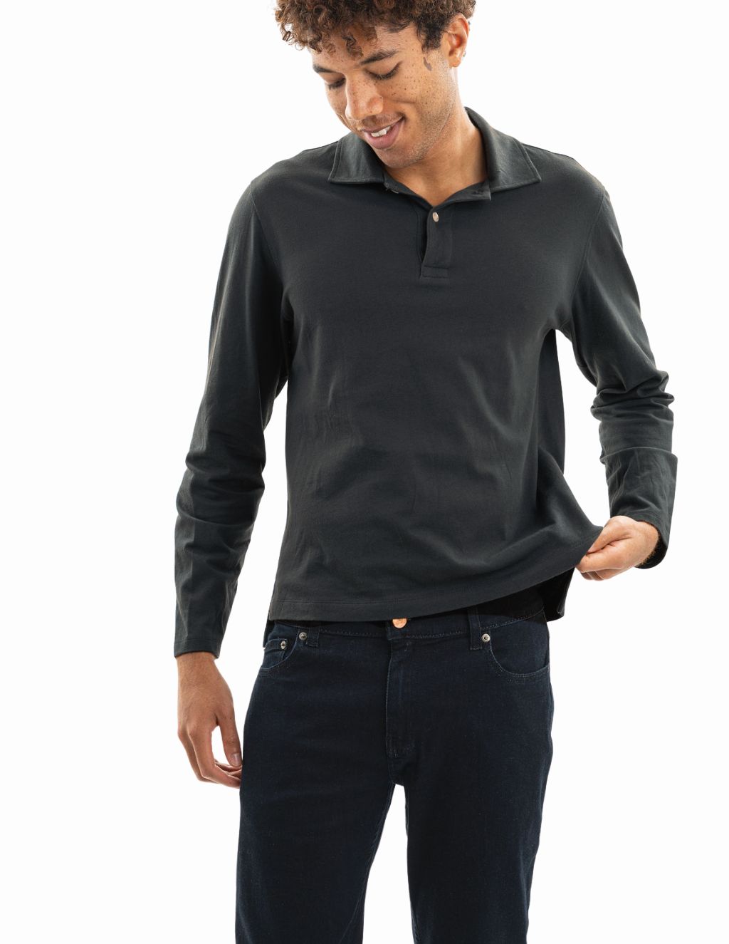 Man wearing dark long-sleeve polo shirt and dark jeans, pulling the hem to show the shirt's fit