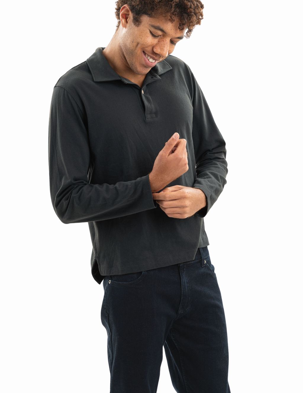 Smiling young man adjusting cuff of dark long-sleeve polo and wearing dark jeans, white studio background.