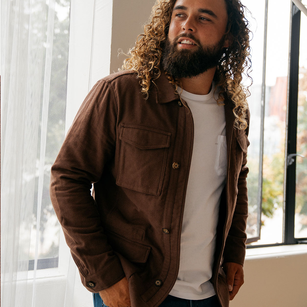 A man with curly hair wearing a brown jacket over a white shirt, standing by a window.