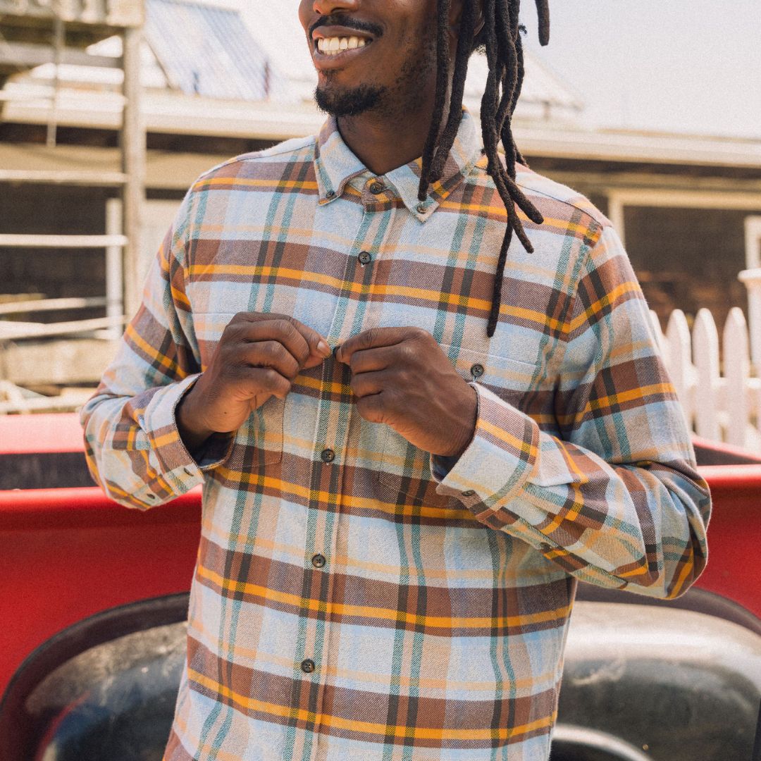Man with dreadlocks smiling while buttoning a brown, blue and yellow plaid shirt by a red truck and white fence.
