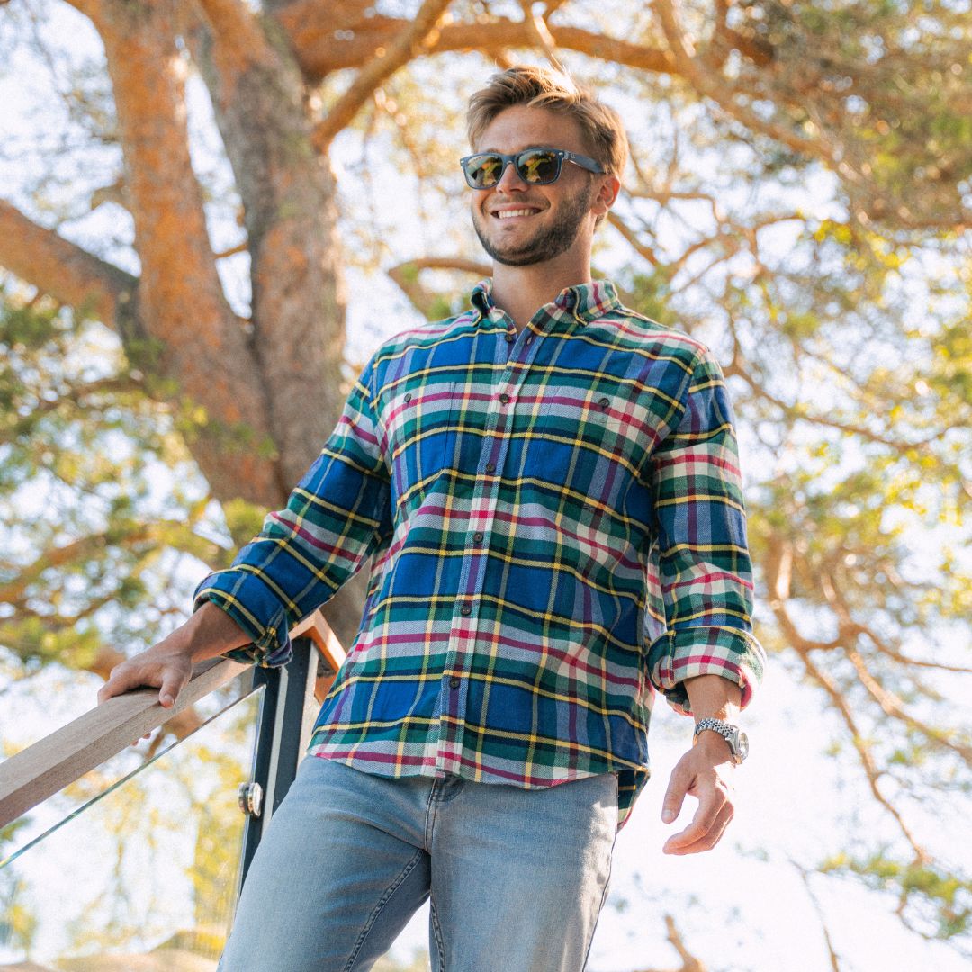 Young man wearing sunglasses and a blue-green plaid shirt, smiling on a balcony railing with sunlit trees behind him.