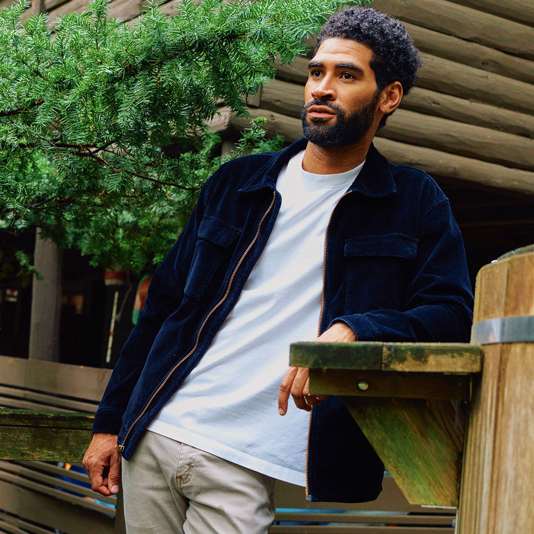 A man wearing a navy corduroy jacket and a white t-shirt, leaning on a wooden railing.