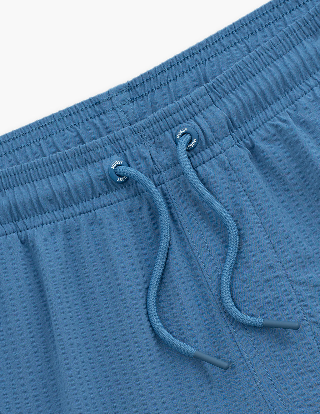 Close-up of Pacifics swim short waistband showing textured blue fabric, elastic drawstring and Mugsy-branded eyelets on white background