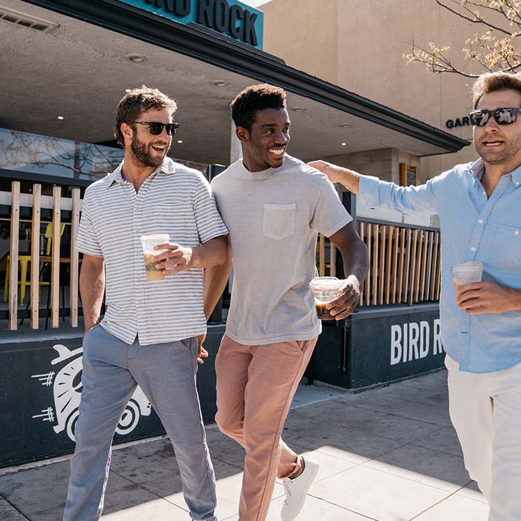 Three men walking and laughing outside a cafe on a sunny day, wearing casual summer outfits and holding iced drinks