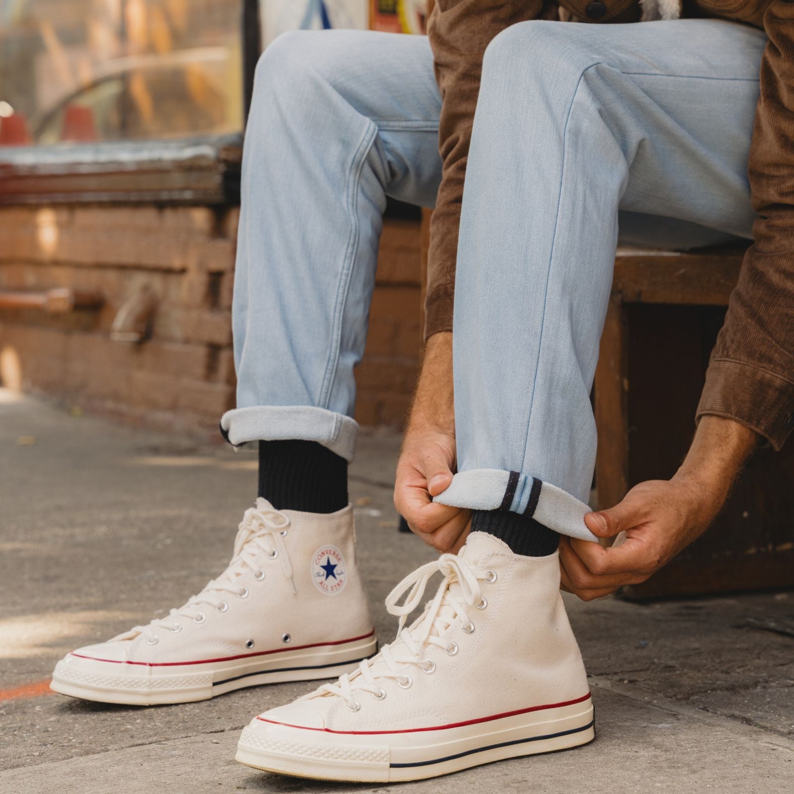 Person rolling cuff of light jeans to reveal black socks and off-white high-top sneakers with CONVERSE ALL STAR patch