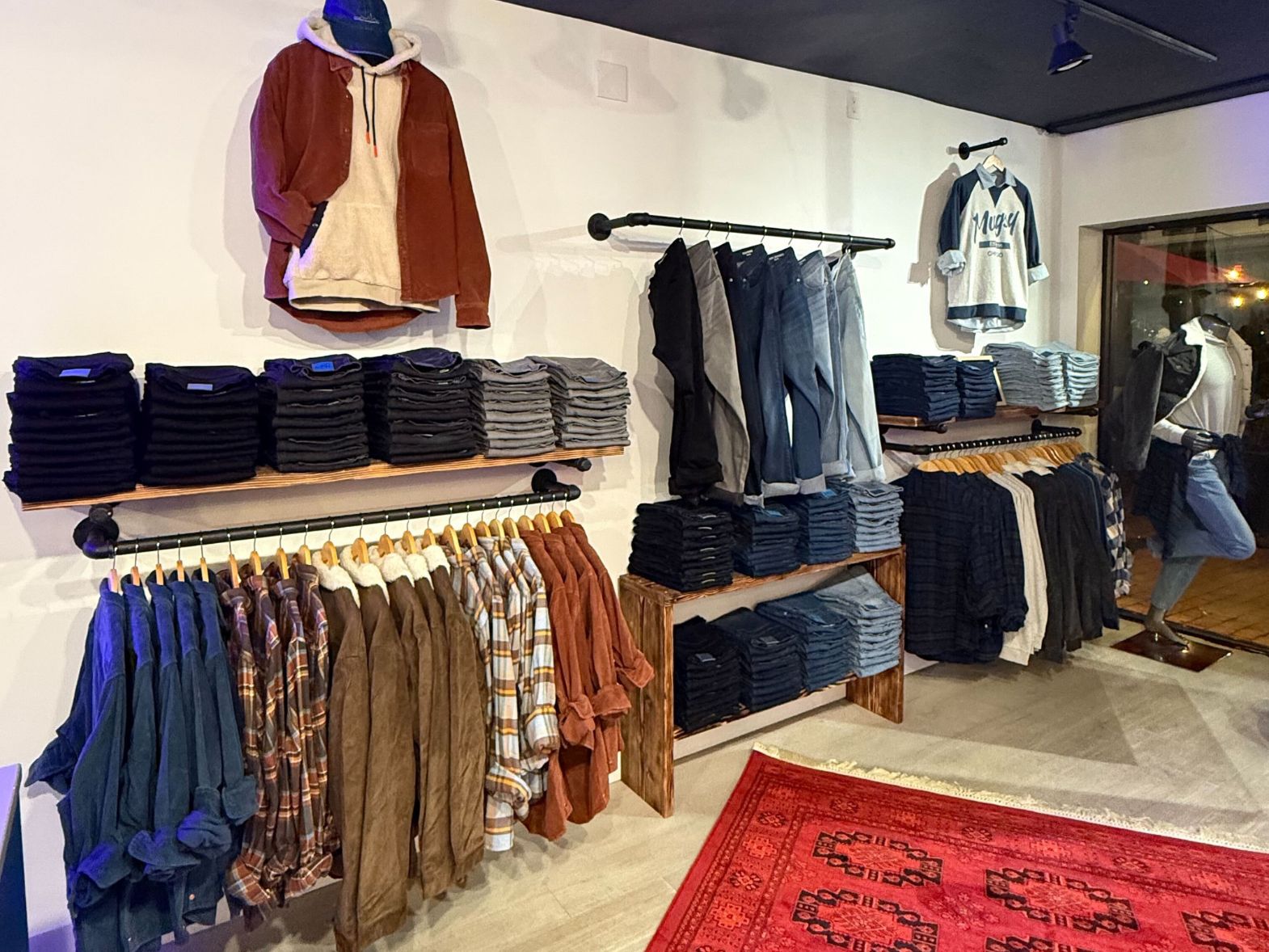 Boutique clothing display with folded jeans, hanging jackets and flannels on wooden racks, mannequin wearing outfit.