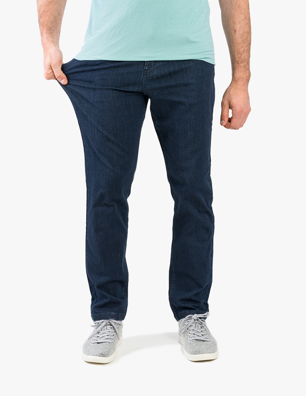Model wearing dark blue casual stretch jeans, pulling left thigh to show fit; mint T-shirt hem and gray sneakers visible