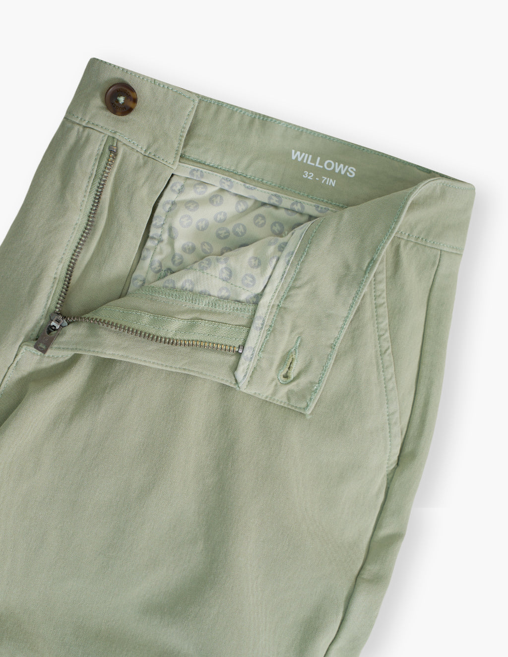 Close-up of Willows chino shorts waistband and zipper showing sage green fabric, printed pocket lining, and button closure on white background