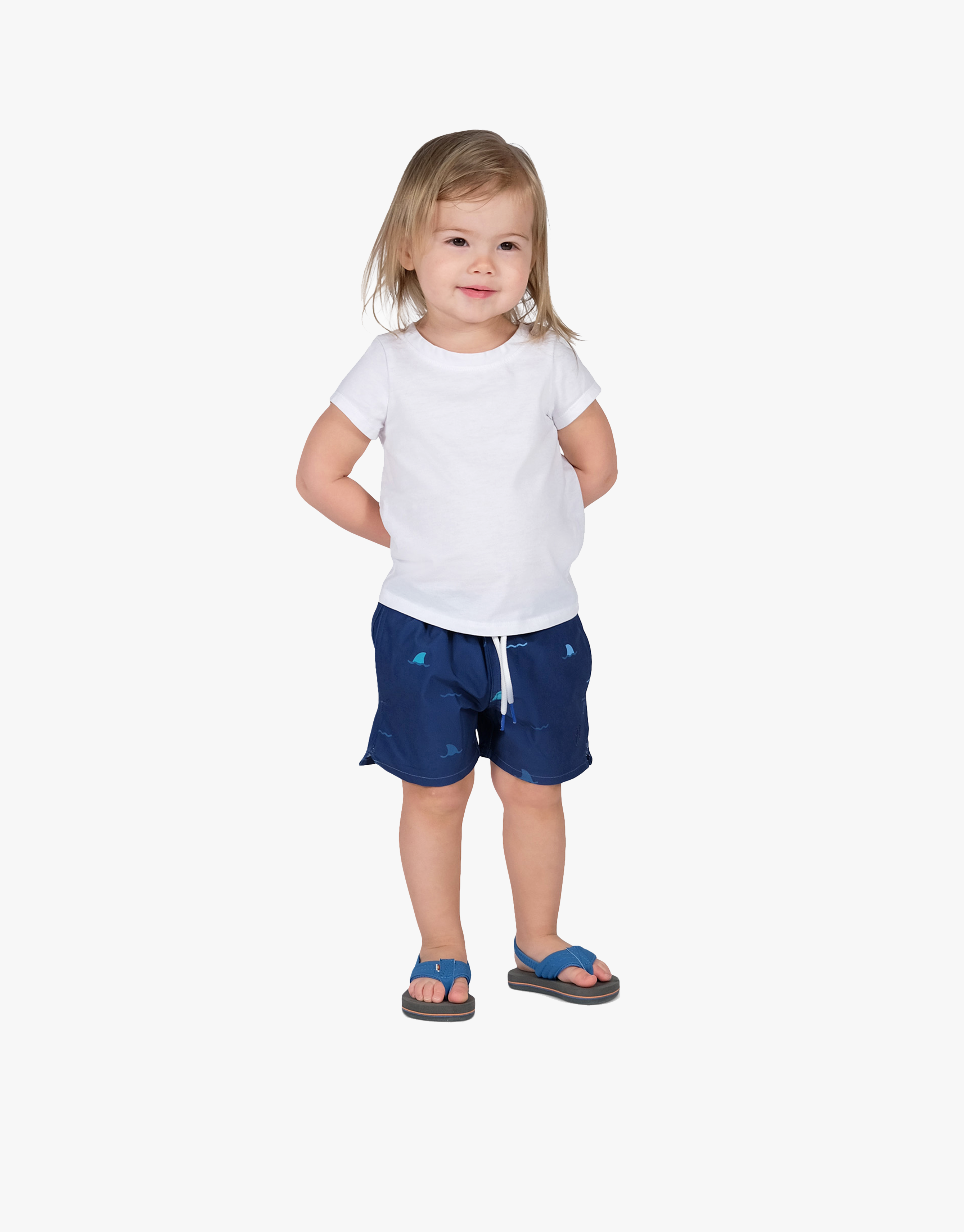 A child wearing navy swim shorts with shark patterns, paired with a white shirt and sandals.